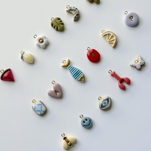 May include: A collection of colorful charms with gold-tone loops. The charms include a cactus, a crescent moon, a leaf, a flower, a lemon slice, a red tomato, a fish, a heart, a lobster, a butterfly, a blue patterned square, a smiley face, and a bee.