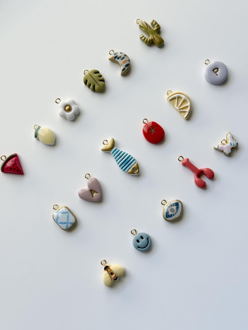 May include: A collection of colorful charms with gold-tone loops. The charms include a cactus, a crescent moon, a leaf, a flower, a lemon slice, a red tomato, a fish, a heart, a lobster, a butterfly, a blue patterned square, a smiley face, and a bee.