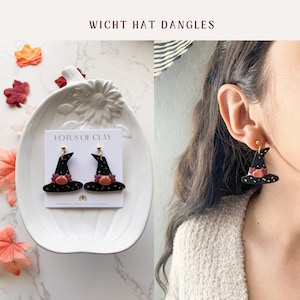 May include: A pair of black witch hat dangle earrings with a gold glitter finish and a small orange pumpkin accent. The earrings are displayed on a white plate with a card that reads "LOTUS OF CLAY HANDMADE JEWELRY & ACCESSORIES".