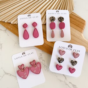 May include: Four sets of pink polymer clay earrings on white cards with the text "LOTUS OF CLAY". The earrings are in the shapes of hearts, teardrops, and squares. Some of the earrings have a leopard print design.