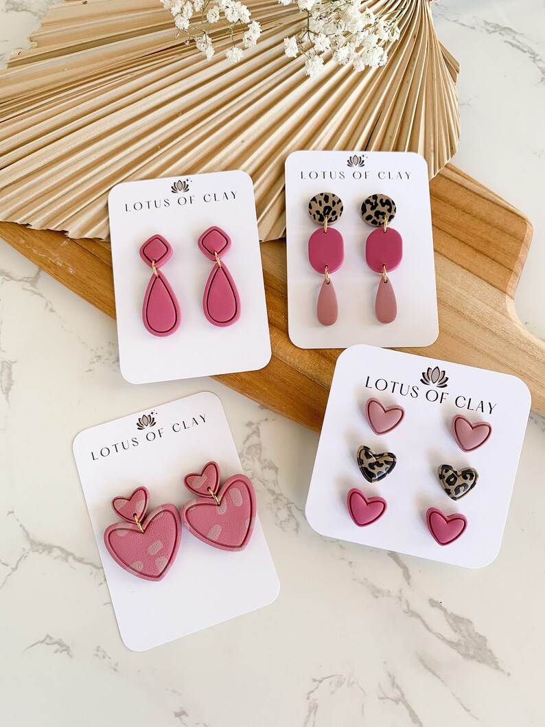 May include: Four sets of pink polymer clay earrings on white cards with the text "LOTUS OF CLAY". The earrings are in the shapes of hearts, teardrops, and squares. Some of the earrings have a leopard print design.