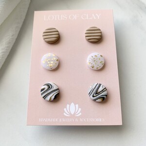 May include: Set of six clay stud earrings. The earrings are in three different designs: brown with horizontal lines, white with gold flecks, and white with black marbling. The earrings are displayed on a pink card with the text "LOTUS OF CLAY HANDMADE JEWELRY & ACCESSORIES".
