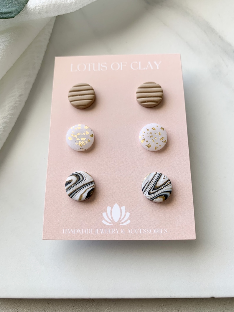 May include: Set of six clay stud earrings. The earrings are in three different designs: brown with horizontal lines, white with gold flecks, and white with black marbling. The earrings are displayed on a pink card with the text "LOTUS OF CLAY HANDMADE JEWELRY & ACCESSORIES".