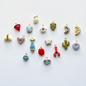 May include: A collection of colorful charms in various shapes, including a watermelon slice, a lemon, a flower, a leaf, a crescent moon, a bee, a heart with the letter "A", a fish, a butterfly, a cactus, a smiley face, a red tomato, a lobster, an eye, and a charm with the letter "P".