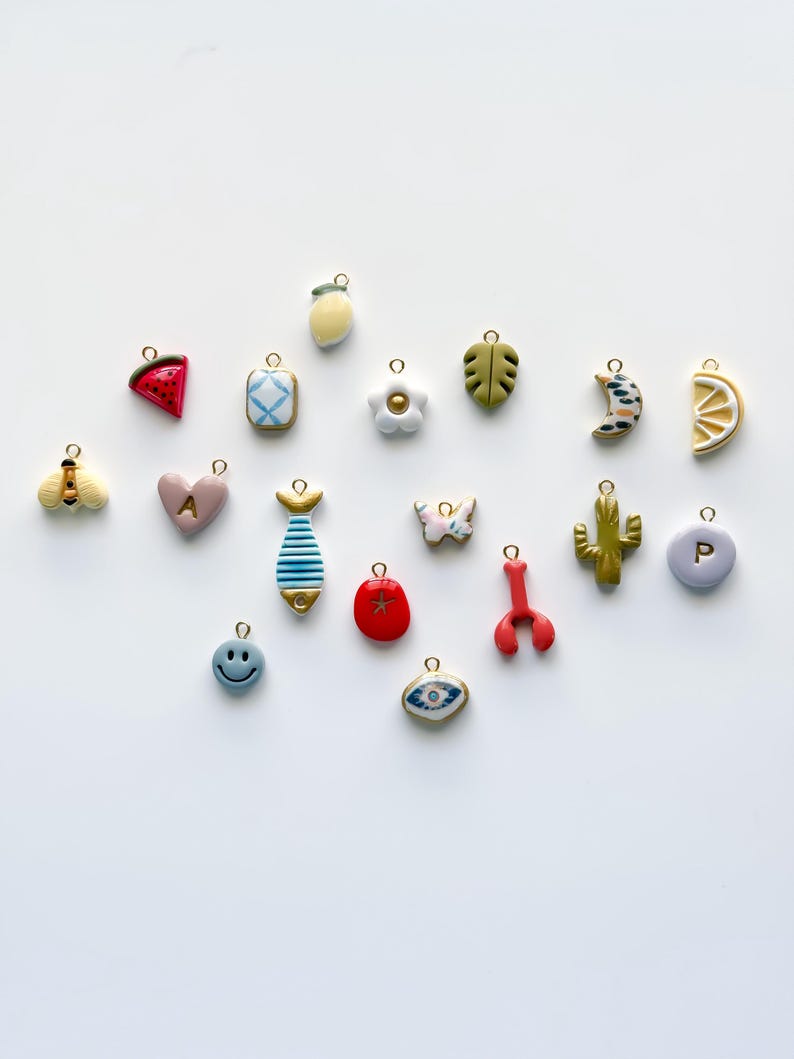 May include: A collection of colorful charms in various shapes, including a watermelon slice, a lemon, a flower, a leaf, a crescent moon, a bee, a heart with the letter "A", a fish, a butterfly, a cactus, a smiley face, a red tomato, a lobster, an eye, and a charm with the letter "P".