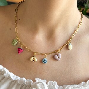 May include: Gold-toned chain necklace with colorful charms. Charms include a watermelon slice, bee, smiley face, heart, leaf, and lemon. The necklace is worn on a person wearing a white top.