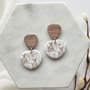 May include: A pair of dangle earrings with a white background featuring a repeating pattern of brown bunnies and floral accents. The top portion of each earring is a brown wood-like material.