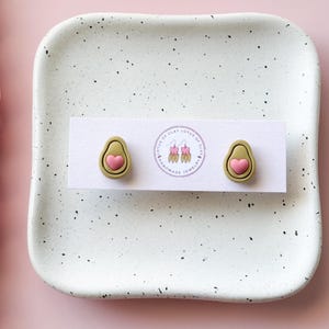 May include: A pair of avocado-shaped stud earrings with a pink heart detail, displayed on a white card. The earrings are olive green with a pink heart center. The card rests on a speckled white ceramic tray.