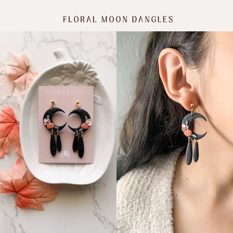 May include: Black crescent moon dangle earrings with small orange pumpkins and pink flowers. The earrings have black teardrop dangles and gold accents. The earrings are displayed on a white plate with a pink card that says "Lotus of Clay" and "Handmade Jewelry & Accessories".