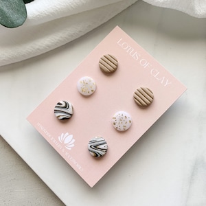May include: A set of six clay earrings in a variety of patterns, including white with gold flecks, black and white marble, and brown with a striped texture. The earrings are displayed on a pink card with the text "Lotus of Clay" and "Handmade Jewelry & Accessories".