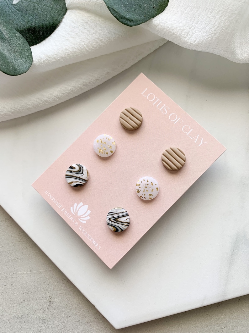 May include: A set of six clay earrings in a variety of patterns, including white with gold flecks, black and white marble, and brown with a striped texture. The earrings are displayed on a pink card with the text "Lotus of Clay" and "Handmade Jewelry & Accessories".
