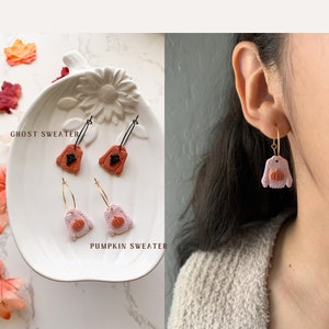 May include: Two pairs of dangle earrings featuring a sweater design. One pair is orange with a black ghost design and the text "Ghost Sweater". The other pair is pink with a small orange pumpkin design and the text "Pumpkin Sweater".