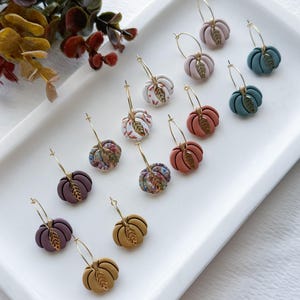 Könnte beinhalten: Eine Sammlung handgefertigter kürbisförmiger Ohrringe in verschiedenen Farben, darunter Rosa, Orange und Grün, auf einem weißen Tablett präsentiert. Jeder Ohrring hat einen goldfarbenen Reifen und ein goldenes Blatt-Akzent, perfekt für den Herbst.