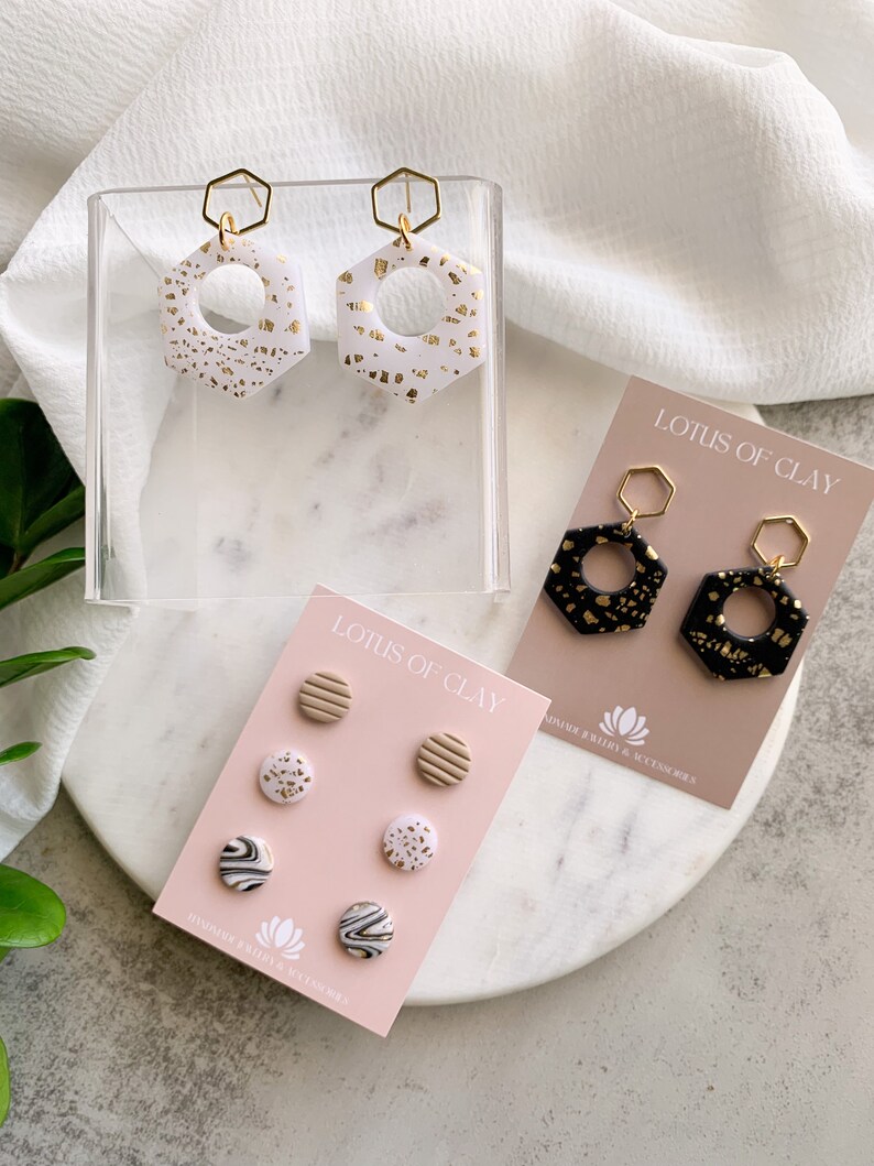 May include: A set of three pairs of handmade polymer clay earrings. The first pair is white with gold flecks, the second pair is black with gold flecks, and the third pair is a set of four small stud earrings with various patterns in white, black, and gold.