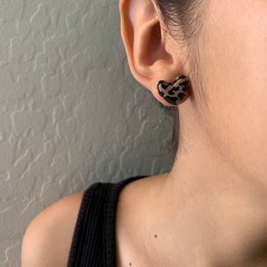 May include: A pair of heart-shaped earrings with a leopard print design. The earrings are gold-toned and have a black and brown leopard print.