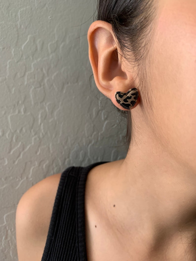 May include: A pair of heart-shaped earrings with a leopard print design. The earrings are gold-toned and have a black and brown leopard print.