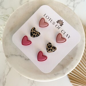 May include: A set of six heart-shaped earrings on a white card. The earrings are pink and have a leopard print design. The card has the text "LOTUS OF CLAY" printed on it.