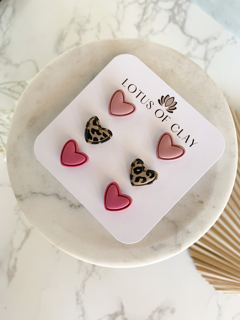 May include: A set of six heart-shaped earrings on a white card. The earrings are pink and have a leopard print design. The card has the text "LOTUS OF CLAY" printed on it.