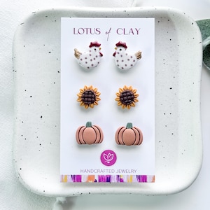 May include: Three pairs of stud earrings on a white display card labeled LOTUS of CLAY. The top pair features white chickens with brown spots and red combs. The middle pair shows yellow sunflowers with brown centers. The bottom pair depicts pink pumpkins.