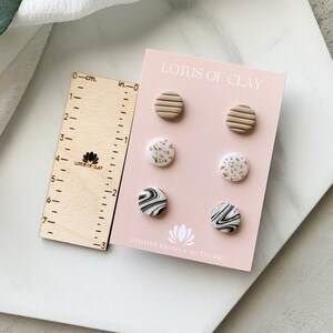May include: A set of six handmade polymer clay earrings. The earrings are in three different designs: striped, speckled, and marbled. The earrings are all in shades of white, black, and gold. The earrings are displayed on a pink card with the text "LOTUS OF CLAY" and "HANDMADE JEWLRY & ACCESSORIES".