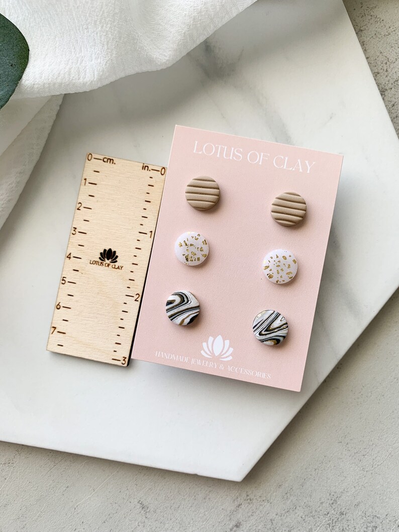 May include: A set of six handmade polymer clay earrings. The earrings are in three different designs: striped, speckled, and marbled. The earrings are all in shades of white, black, and gold. The earrings are displayed on a pink card with the text "LOTUS OF CLAY" and "HANDMADE JEWLRY & ACCESSORIES".