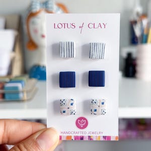 May include: A card displaying six square stud earrings. The earrings feature various designs: white with blue stripes, solid navy blue, and patterned with blue and beige floral motifs. The card reads "LOTUS of CLAY" and "HANDCRAFTED JEWELRY".
