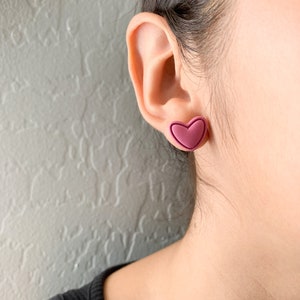 May include: A pair of heart-shaped earrings in a deep pink color. The earrings are made of a smooth, matte material and have a slightly raised edge.