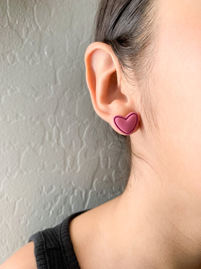 May include: A pair of heart-shaped earrings in a deep pink color. The earrings are made of a smooth, matte material and have a slightly raised edge.