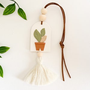 May include: White ceramic car air freshener with a brown leather cord and a white tassel. The ceramic charm features a green potted plant with brown leaves.