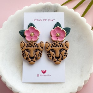 May include: A pair of handmade earrings featuring a cheetah face design with pink flower accents and green leaves. The earrings are displayed on a white card with the text "LOTUS OF CLAY".