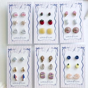 May include: Six display cards labeled Lotus of Clay, each holding three pairs of small stud earrings. The designs include seashells, sailboats, fruit, ice cream cones, striped shirts, hats, geometric tiles, fish, and various abstract shapes in many colors.