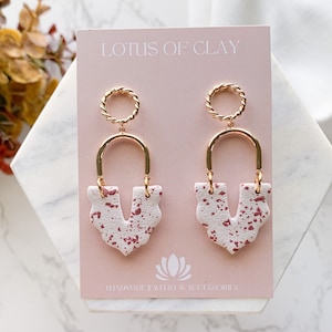 Peut inclure: Une paire de boucles d'oreilles pendantes dorées et blanches avec un motif moucheté rose et blanc. Les boucles d'oreilles présentent un anneau doré en haut et une arche dorée avec un motif moucheté rose et blanc qui pend de celui-ci. Les boucles d'oreilles sont étiquetées "LOTUS OF CLAY" et "HANDMADE JEWELRY & ACCESSORIES".