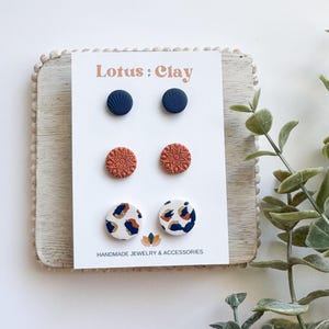 May include: A set of six stud earrings displayed on a wooden card. The earrings are made of polymer clay and feature three different designs: navy blue, rust orange, and white with navy blue and rust orange accents. The card has the text "Lotus Clay" and "Handmade Jewelry & Accessories".