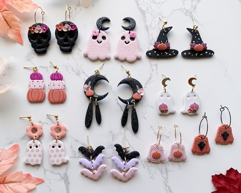 May include: A collection of 18 Halloween-themed polymer clay earrings. The earrings feature various designs, including ghosts, pumpkins, bats, skulls, witches' hats, and moons. The earrings are in shades of pink, black, white, and orange.
