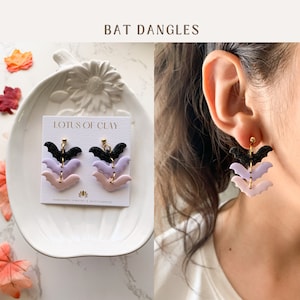 May include: A pair of dangle earrings featuring three layered bats in black, lavender, and pink. The earrings are displayed on a white card with the text "LOTUS OF CLAY HANDMADE JEWELRY & ACCESSORIES".