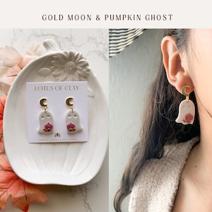 May include: A pair of white ghost earrings with a gold crescent moon and a pink pumpkin. The earrings are on a white card with the text "LOTUS OF CLAY" and "HANDMADE JEWELRY & ACCESSORIES".