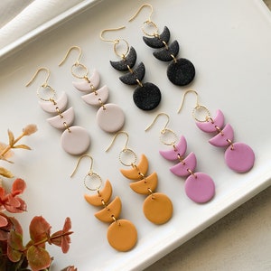 Può includere: Un set di cinque paia di orecchini pendenti con tre forme a mezzaluna e un cerchio. Gli orecchini sono realizzati in argilla polimerica e sono disponibili in una varietà di colori, tra cui nero, bianco, rosa, giallo e arancione.