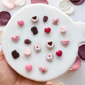 May include: A white ceramic plate with a variety of pink and red heart-shaped and other Valentine's Day themed polymer clay charms. The charms include hearts, cookies, strawberries, and chocolate.