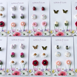 May include: A collection of handmade stud earrings featuring floral, insect, and abstract designs. The earrings are displayed on cards with the "LOTUS of CLAY" logo and a floral background. The earrings include daisies, butterflies, and ladybugs.