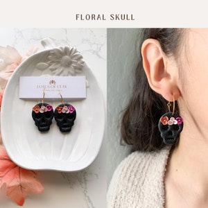 May include: Black skull earrings with pink and white flower accents. The earrings are on a white plate with a business card that says "Lotus of Clay".