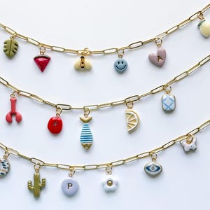 May include: Three gold chain necklaces with colorful charms. Charms include a leaf, a red gem, a bee, a smiley face, a heart with an "A", a lobster claw, a tomato, a fish, a lemon slice, a square, a crescent moon, a cactus, a letter "P", a flower, an eye, a butterfly, and a lemon.
