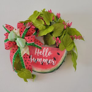May include: A watermelon-shaped wreath with green leaves and pink flowers. The watermelon has the words "Hello Summer" written on it in white.