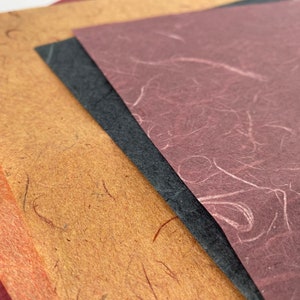 Peut inclure: Une pile de feuilles de papier faites à la main de différentes couleurs, dont orange, marron, noir et bordeaux. Le papier a une surface texturée avec des fibres visibles, adapté aux travaux manuels et à la papeterie.
