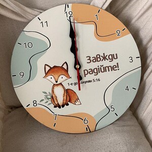 Puede incluir: Un reloj de pared redondo con una ilustración de un zorro de dibujos animados. La esfera del reloj es blanca con números y manecillas negras. El diseño incluye detalles en naranja y azul, y texto en idioma extranjero.