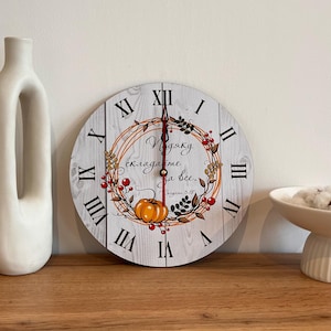 Puede incluir: Un reloj de madera blanco con números romanos y un diseño de otoño. La esfera del reloj presenta una corona de hojas, bayas y calabazas con el texto "Estoy agradecido por todo." en ruso.