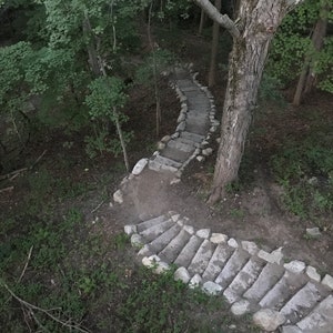 May include: Stone steps winding up a wooded hillside. The steps are made of flat, rectangular stones with rounded edges. The steps are surrounded by green foliage and trees.