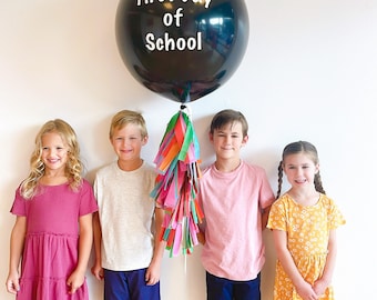 May include: Four children stand in front of a white wall with a black balloon that says "First Day of School". The balloon is tied to a colourful tassel. The children are wearing casual clothing and smiling.