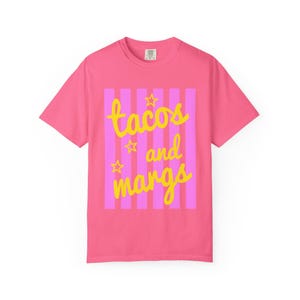 May include: A pink t-shirt featuring the words "tacos and margs" in yellow script, set against a pink and purple striped background. The shirt has a crew neck and short sleeves. The design includes star accents.