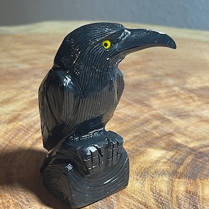 May include: A black obsidian raven sculpture with a yellow eye, perched on a carved wooden base.