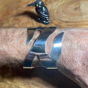 May include: A silver bracelet with a wavy design. The bracelet is on a person's wrist.